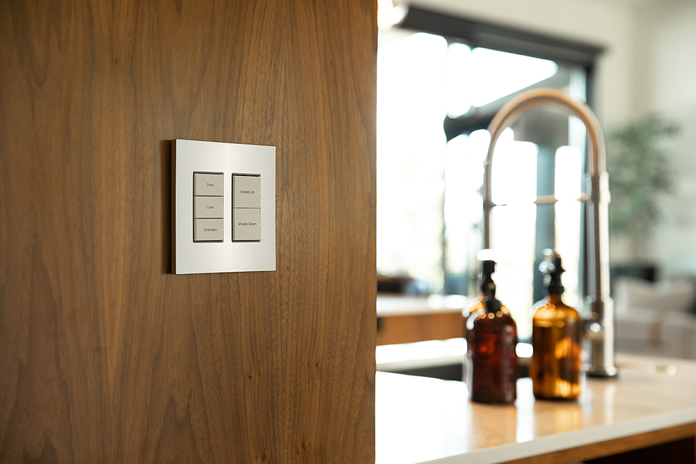 A pair of lighting control keypads on a wood wall with a kitchen in the background. The left keypad has 3 buttons and the right has 2.